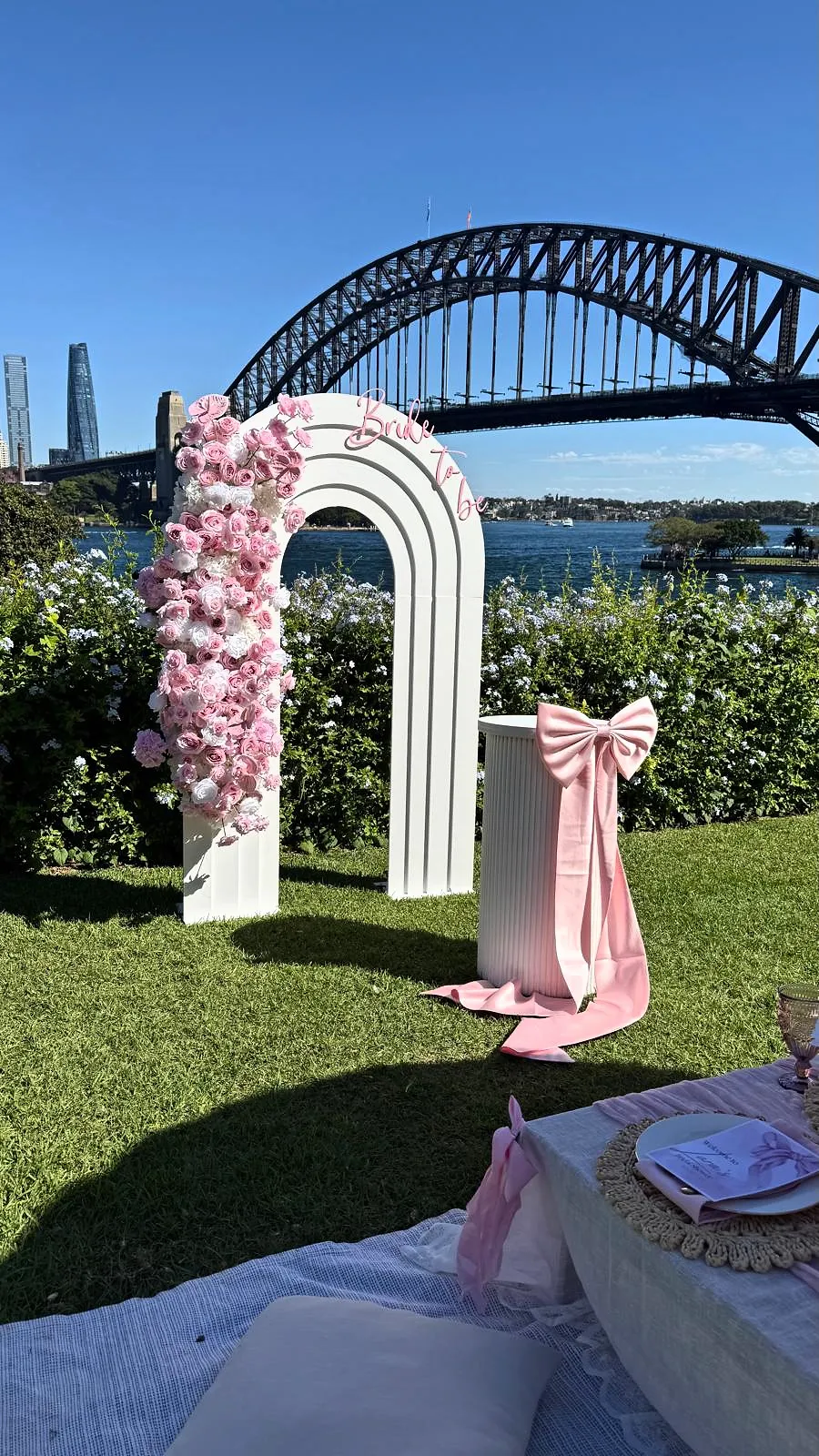 Pink floral arch with a satin bow beside a picnic, with Sydney Harbour Bridge behind