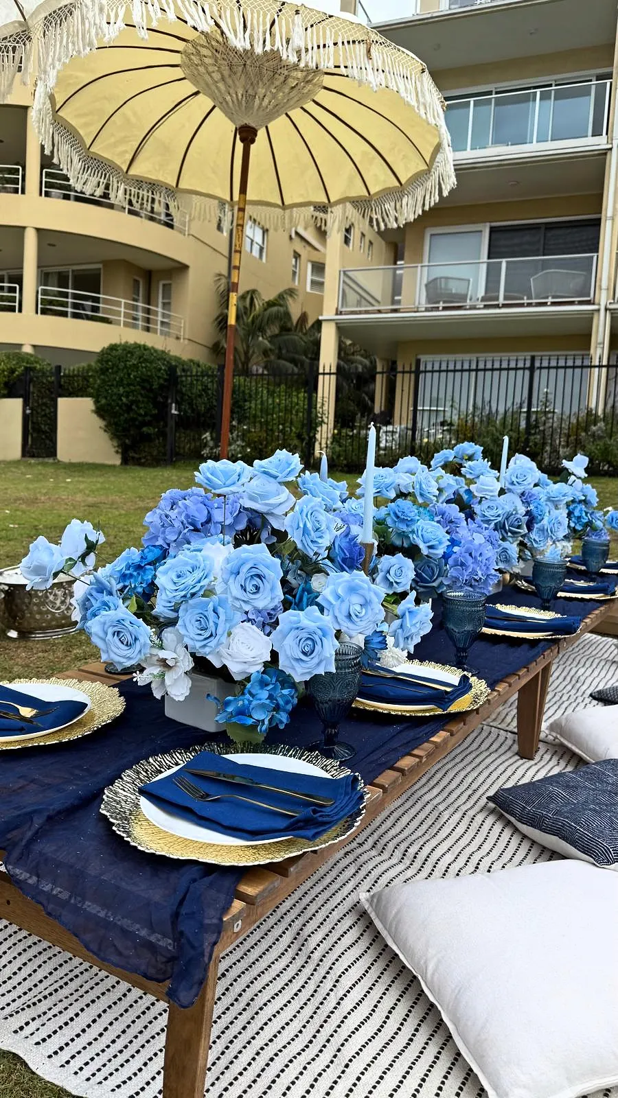 Low picnic table with blue rose centrepieces, navy linen and gold chargers under a fringed umbrella