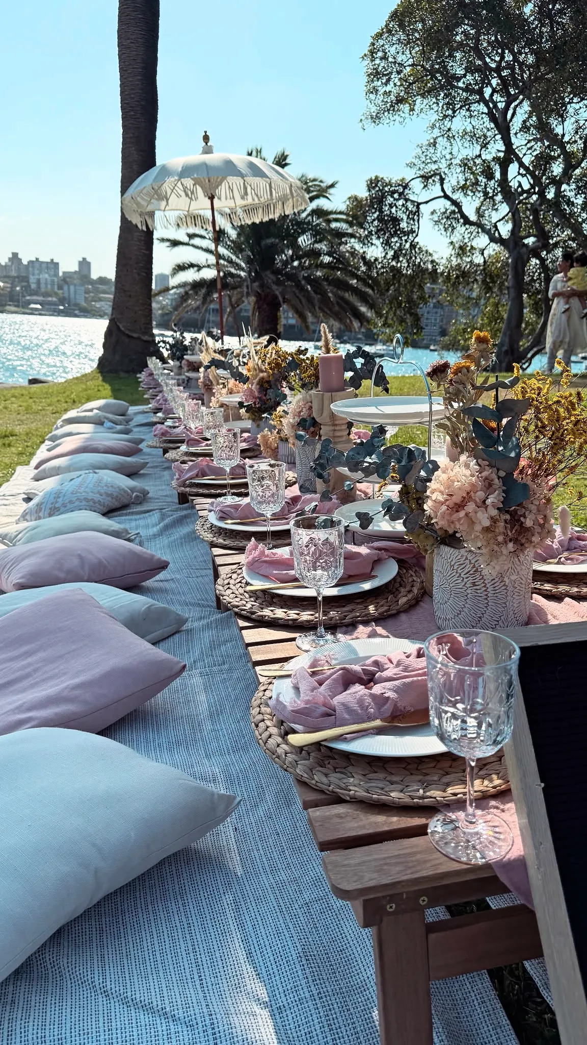Long picnic table with blush cushions, dried florals and fringed umbrellas on a harbourside lawn