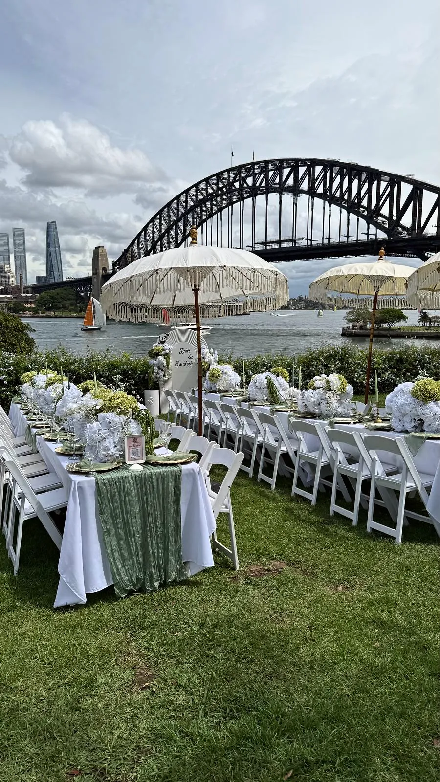 Long tables with sage linen, white chairs and fringed umbrellas on the lawn below Sydney Harbour Bridge