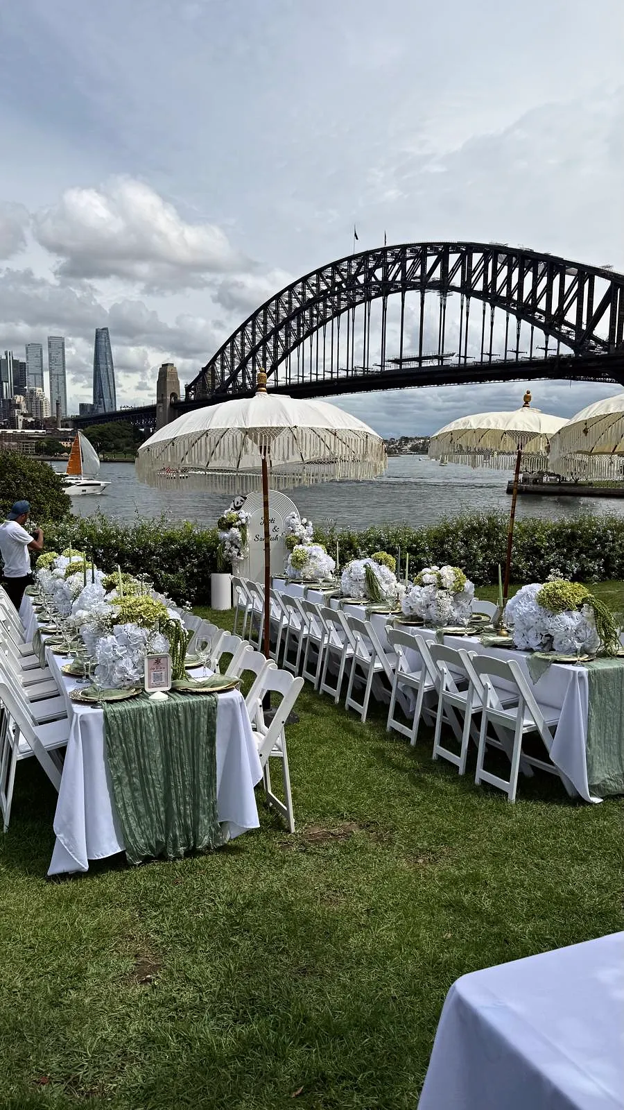 Long tables with sage runners, white chairs and fringed umbrellas beside Sydney Harbour Bridge
