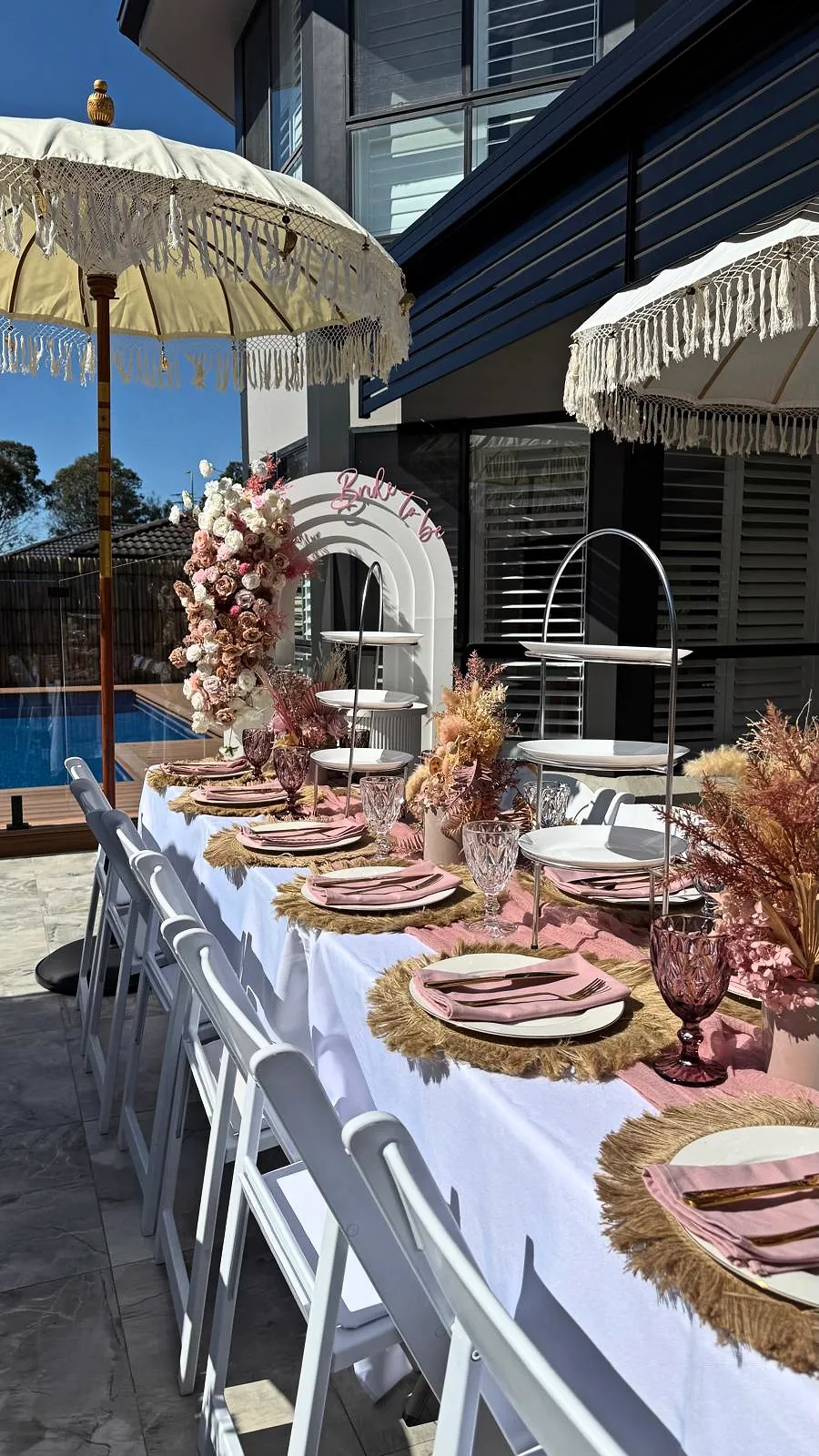 Long table with pink florals, gold chargers and a tiered stand beside a pool under fringed umbrellas