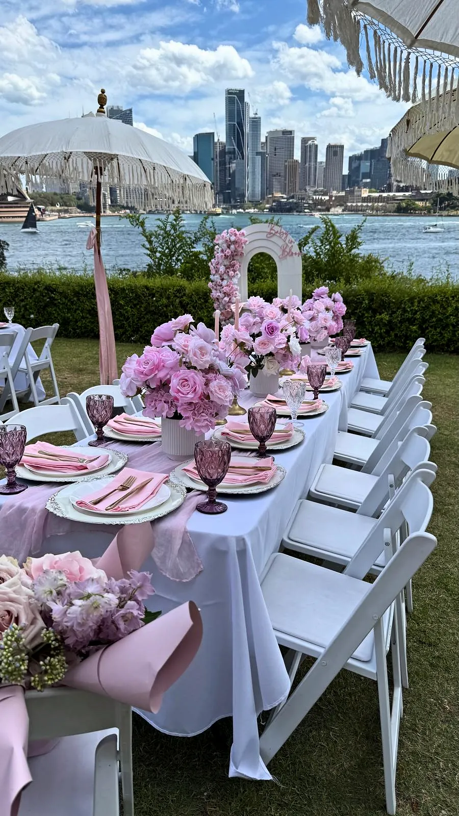 Long table with pink hydrangeas and white chairs on a harbourside lawn, with the city skyline behind