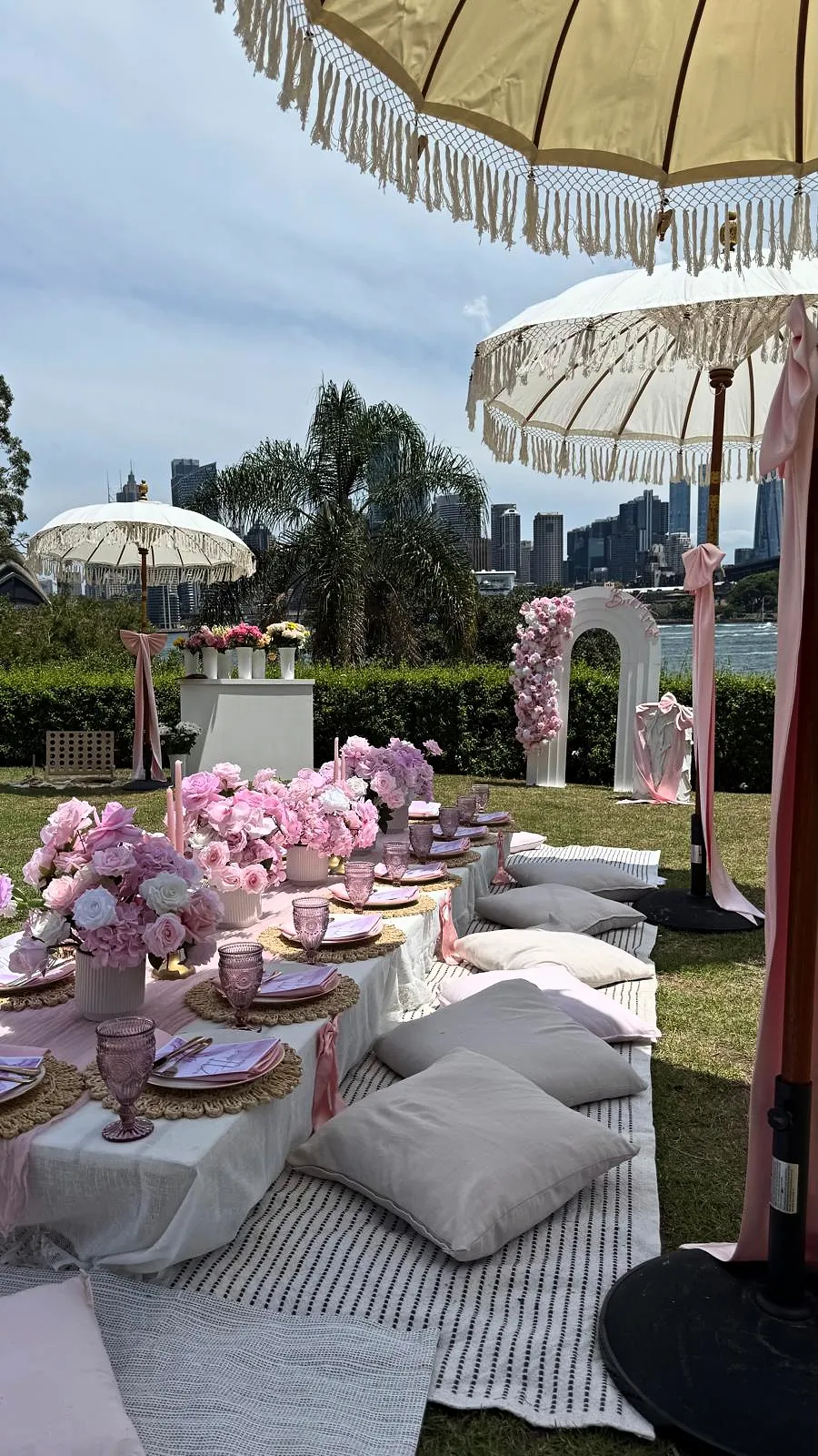 Pink picnic with low tables, fringed umbrellas and a floral arch, with the Sydney skyline across the water