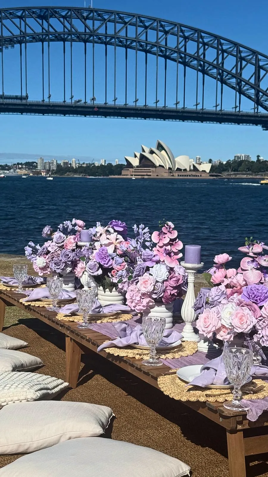 Long low picnic table with pink and lilac florals beside Sydney Harbour, with the Harbour Bridge above and the Opera House across the water