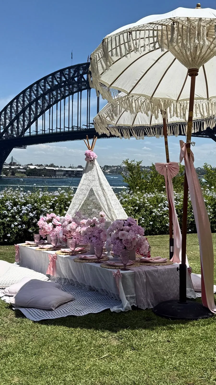 Lace teepee and pink picnic table under fringed umbrellas beside Sydney Harbour Bridge