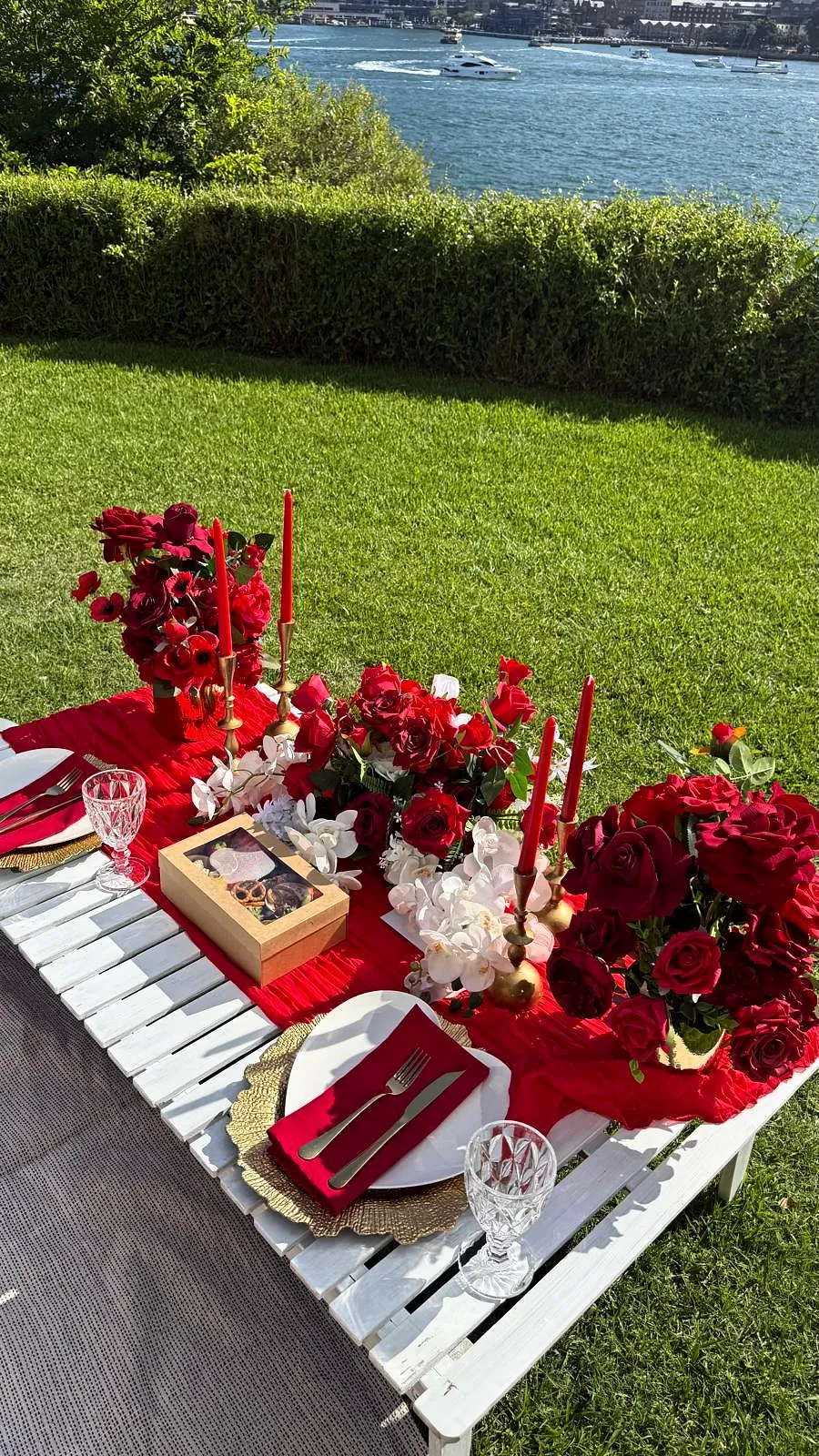 Low white picnic table styled with red roses, red linen and candles beside the water