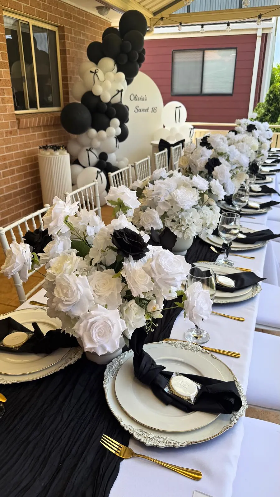 Black and white table with white roses, black napkins and gold-rimmed plates beside a balloon backdrop