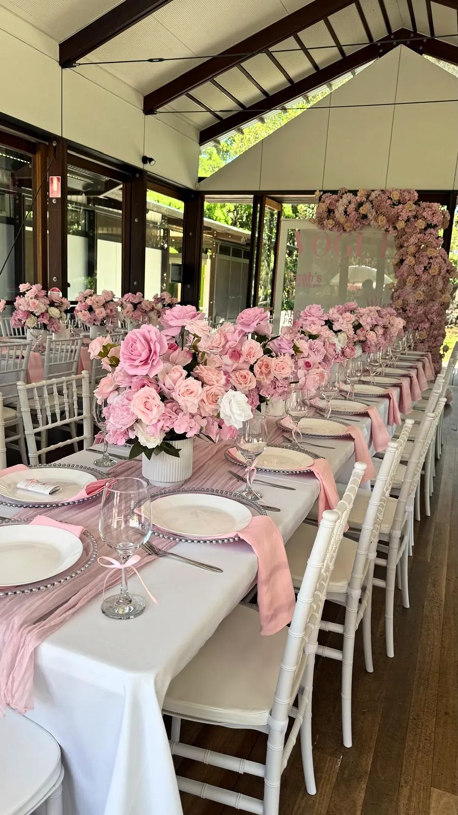 Long table with pink rose centrepieces, pink napkins and white chairs in a glass-walled pavilion